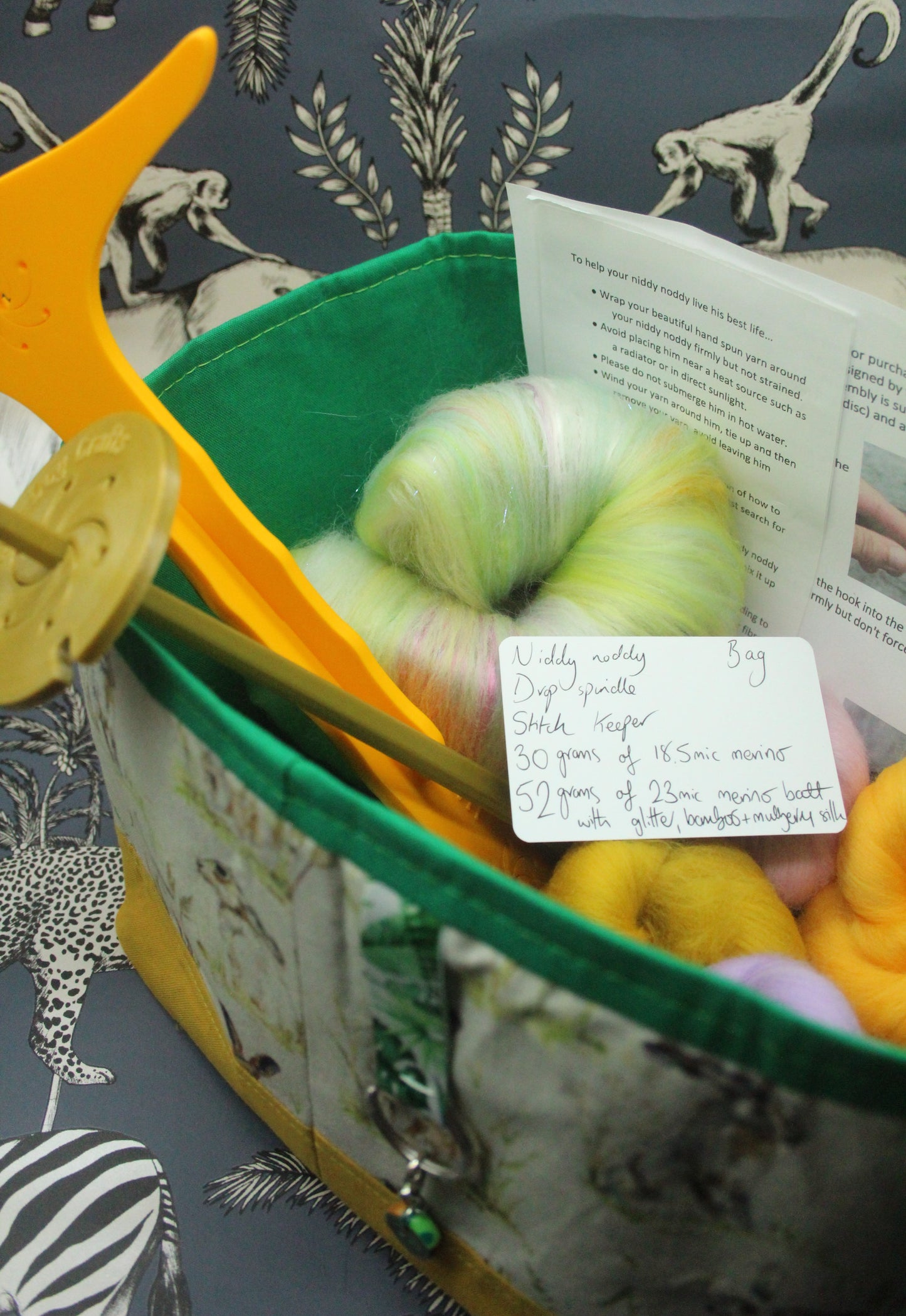 A close-up of a yarn skein bundle inside a patterned fabric basket or bag, with knitting tools and a handwritten note visible. The yarn is variegated in pale green, cream, and soft yellow tones.